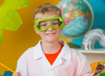A boy in a lab coat and goggles stands in a science-themed room.