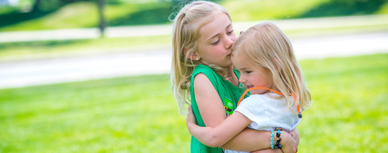 An elementary aged girl hugs a preschool girl.