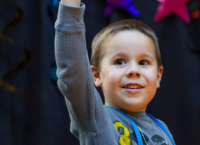 An elementary aged boy raises his hand as he smiles curiously.