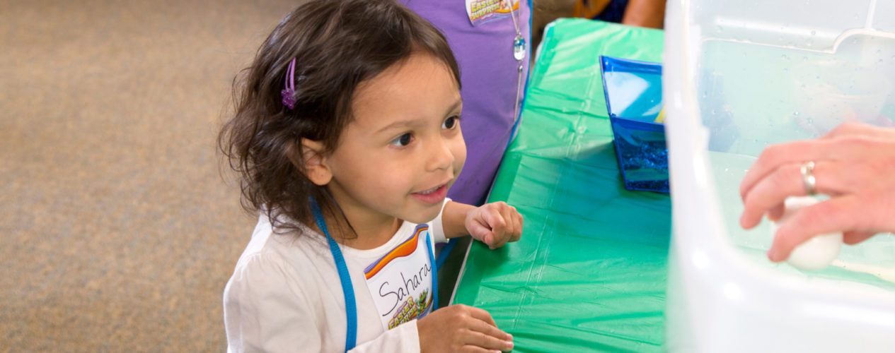 A preschool girl watches the object lesson about Jesus turning water into water in awe.