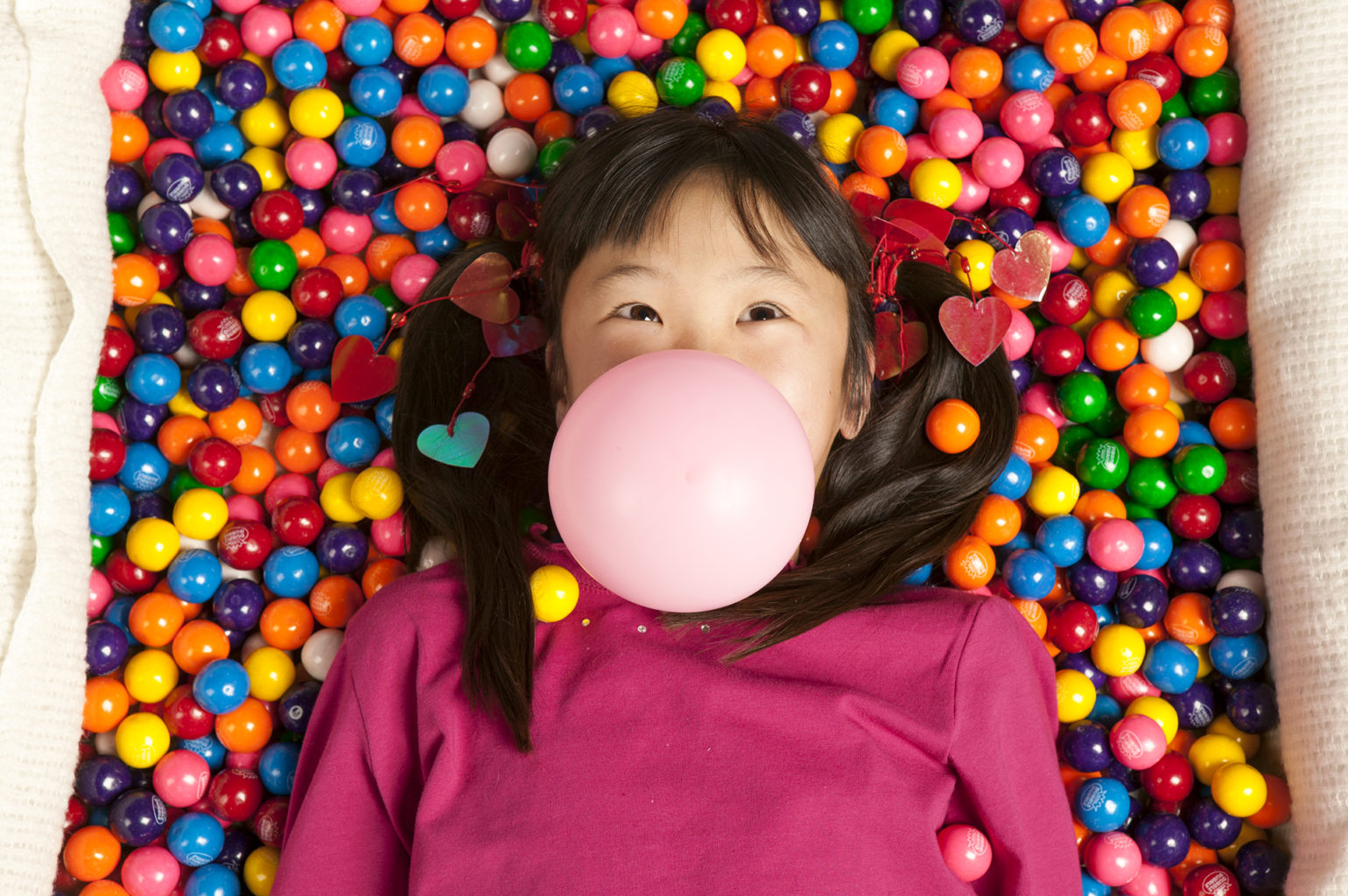 A girl is laying on top of a pile of gum balls. She's blowing a bubble out of chewing gum.