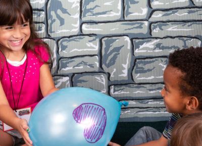 A preschool girl is passing a blue balloon with a purple heart on it to a preschool boy.