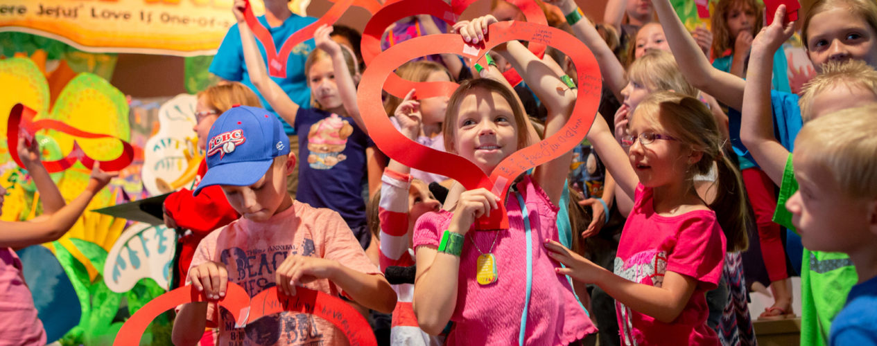 A large group of children are holding out outlines of construction paper hearts around their faces.