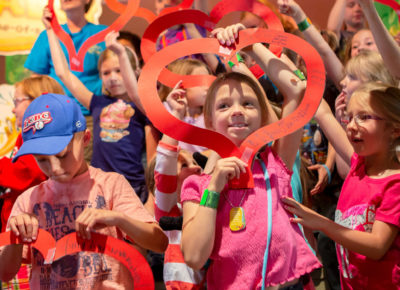 A large group of children are holding out outlines of construction paper hearts around their faces.
