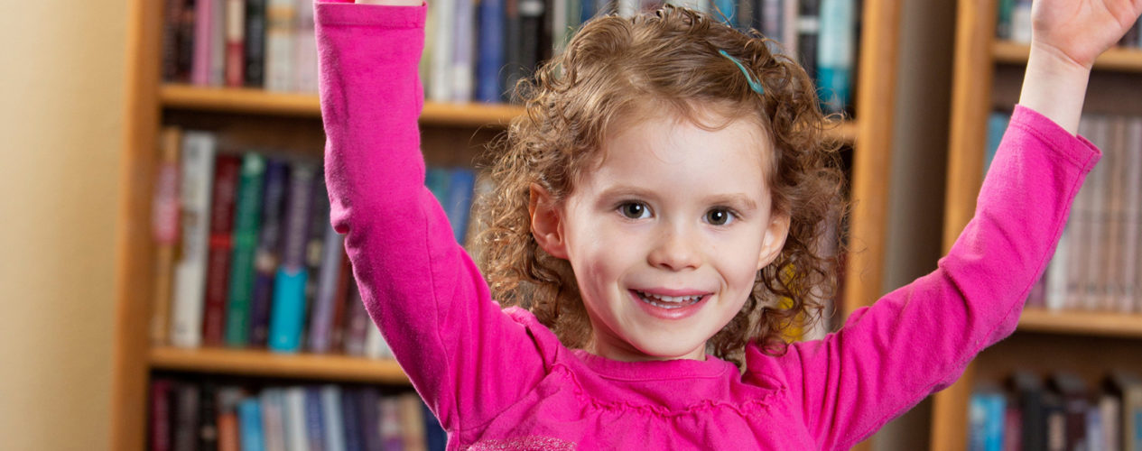 A preschool girl raises her hands and smiles in excitement.