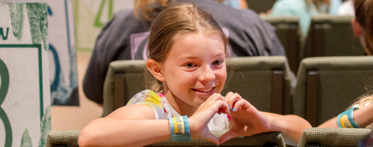 An older elementary girl turns backwards in her chair and makes a heart with her hands.