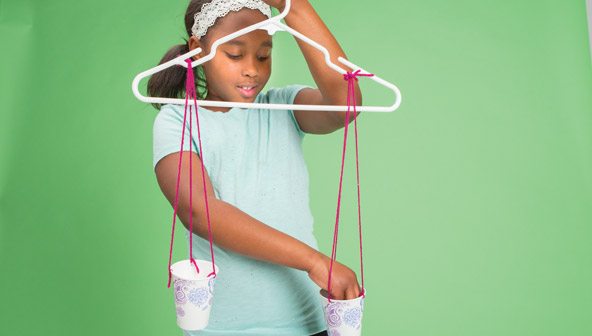 An elementary girl is holding a plastic hanger by the hook. The hanger has two paper cups attached with yarn. She is moving something from one cup to another.
