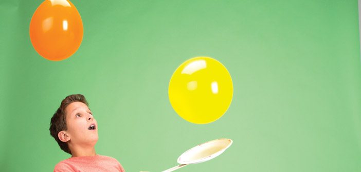A boy is playing with a paint stick and paper plate paddle as he hits two balloons in the air.