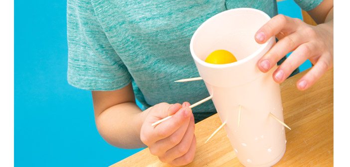 A child is pulling a wooden skewer from a styrofoam cup. There is a yellow ping-pong ball in the cup.