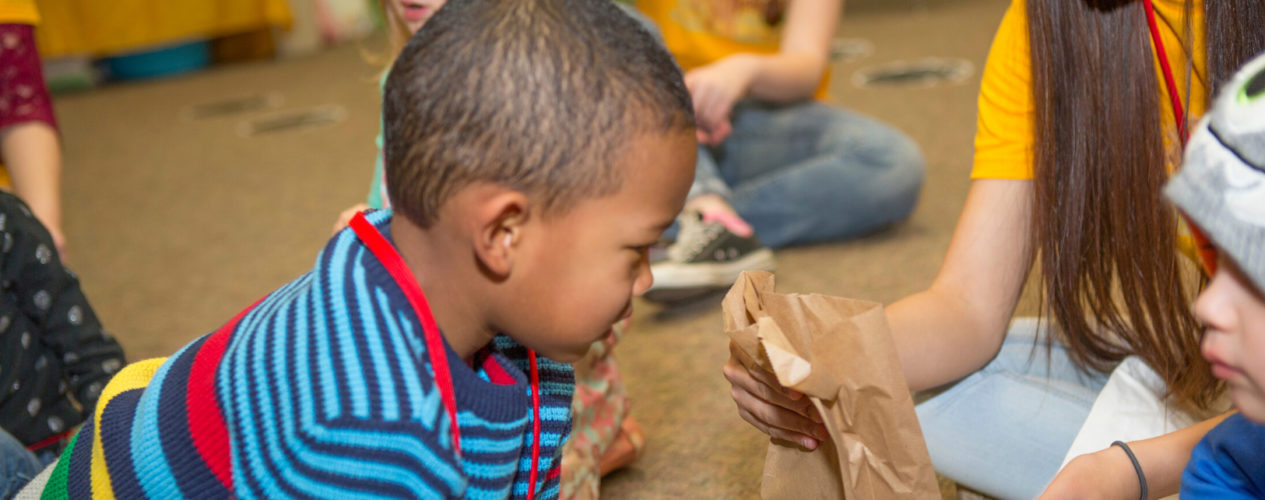 A preschool boy looks inside a brown paper bag.