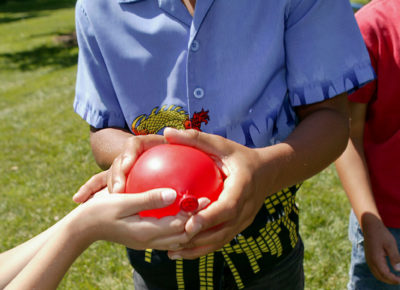 Red water balloon passing between two children's hands. during an Olympic-style family event.