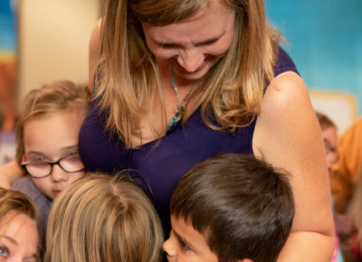 Lady hugging a group of kids. Everyone is smiling.