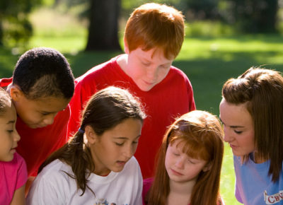 A group of preteens gathered around with an adult volunteer. They are outside gathered around a picnic table, reading the Bible.
