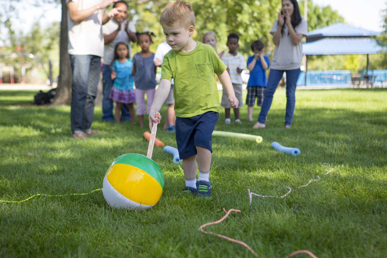 A preschool boy pushing a beach ball with a paint stick across the grass.