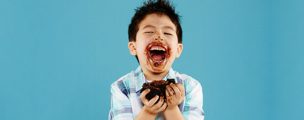 Boy, standing in front of a bright blue background, excitedly has a huge chunk of cake in his hand. His smile is covered in frosting.