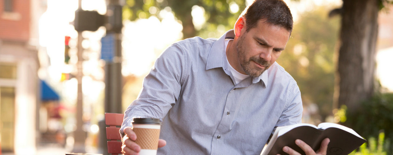 Middle-aged man sits at a an outside table readying his Bible and drinking coffee.