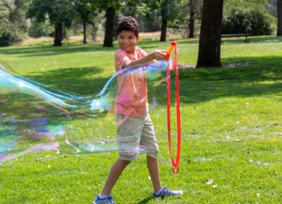 A preteen boy making a huge bubble outside.