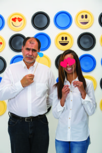 An older adult male and younger female volunteer participate in an emoji photo op. She has heart eyes and he has a tear by his face.