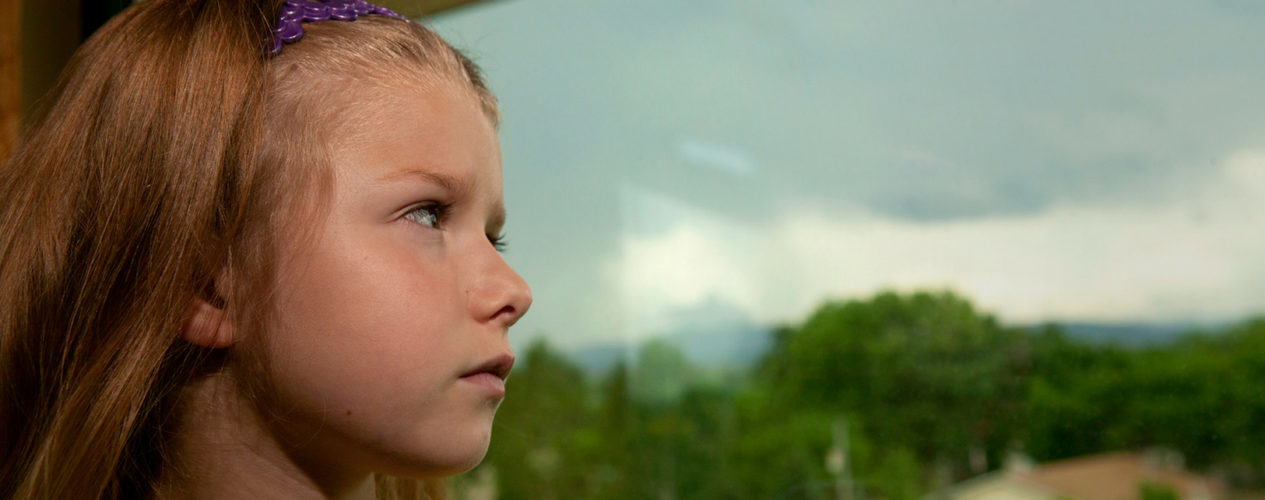 Elementary-aged girl in crisis looks out the window. It's gloomy outside and her brow is furrowed.