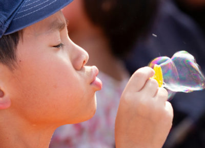 Elementary-aged boy wearing a blue hat faces sideways as he blows a bubble.