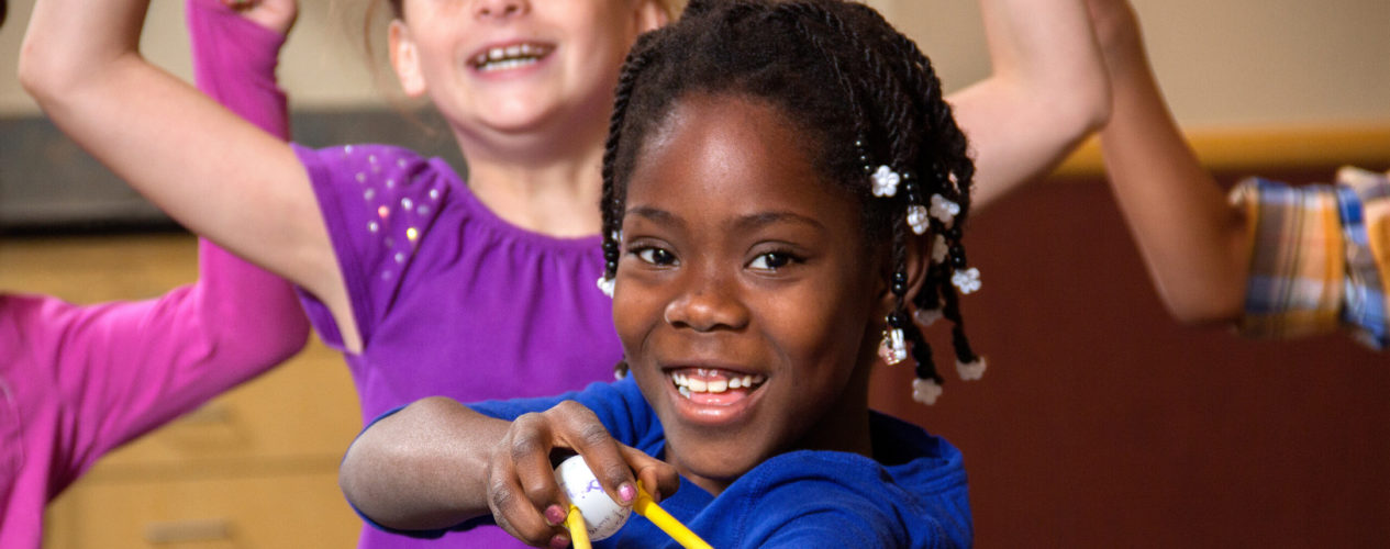 An elementary aged girl pulls back a slingshot with a ping pong ball in it. Other children are cheering for her during the Sunday school game.