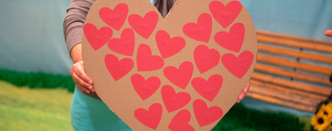 A huge cardboard heart is being held up. On the heart, there are many, small red Post-It-Note hearts.