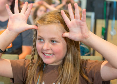 A preteen girl has her hands on the top of her head like antlers. She's engaged in the lesson being taught.