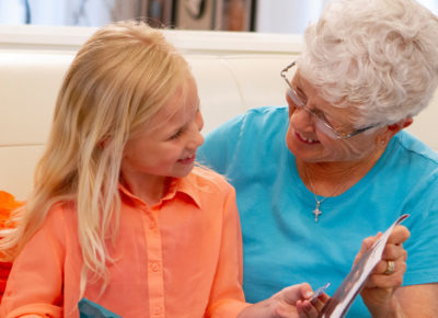 Elementary-aged girl sitting on the couch with her elderly neighbor. They are reading a book together.