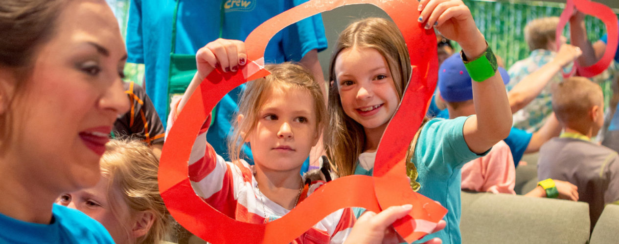 Two elementary-aged girls smiling as they hold a construction paper outline of a heart around their faces.