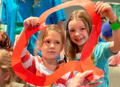 Two elementary-aged girls smiling as they hold a construction paper outline of a heart around their faces.