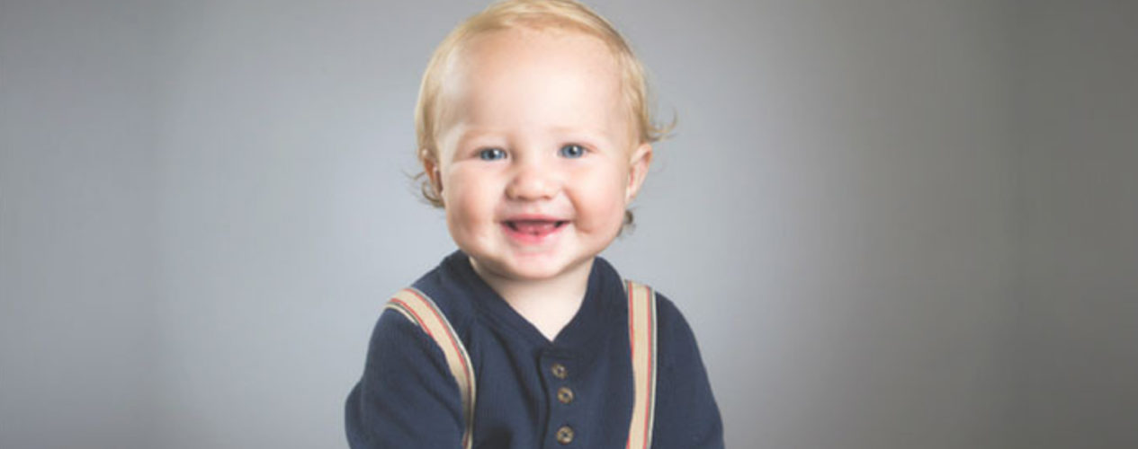 A toddler sitting in front of a gray background smiling. He is wearing suspenders,