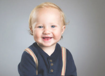 A toddler sitting in front of a gray background smiling. He is wearing suspenders,
