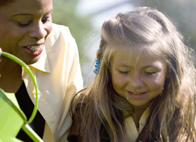 A female volunteer helps an elementary-aged girl water some flowers.