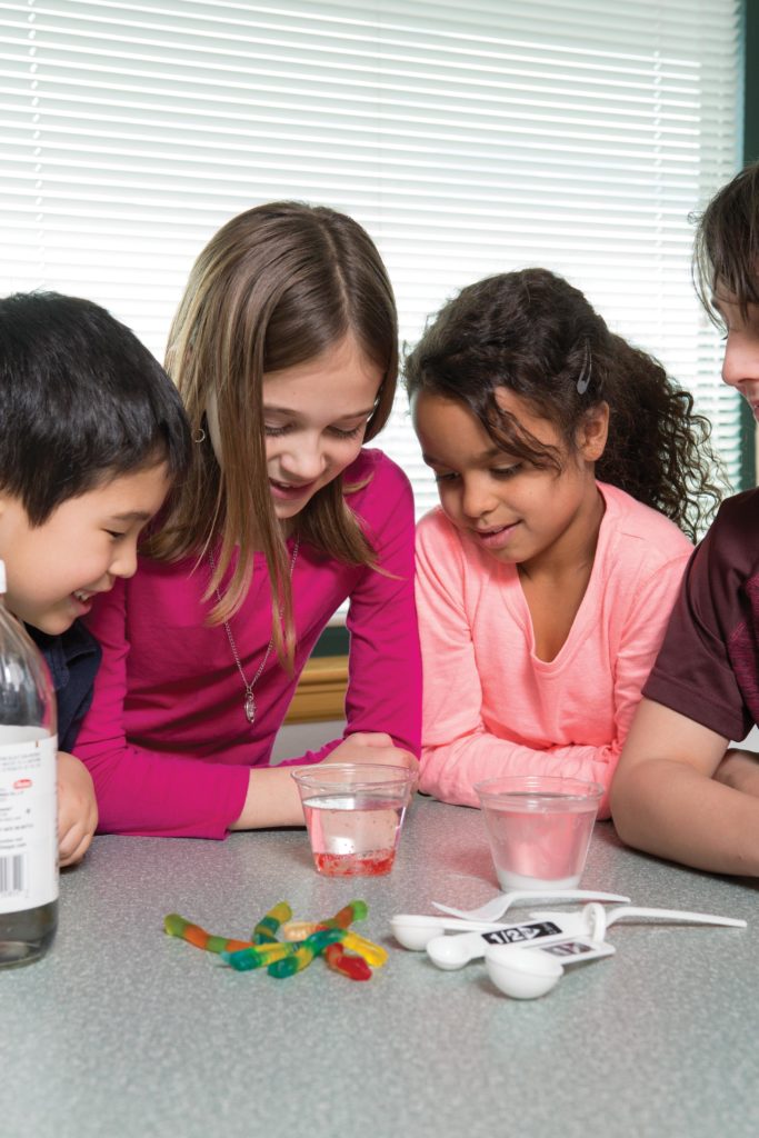 4 children standing around a table looking at a small cup of water with a gummy worm in it.