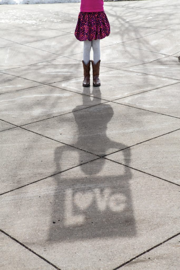 A elementary girl is holding up a cardboard sign with the word "Love" cut out. We see her shadow on the ground with the word "Love" visible.