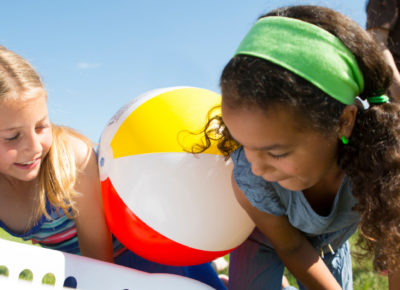 Three elementary-aged girls crawling on the grass. They are trying to get an inflatable beach ball into a laundry basket without using their hands.