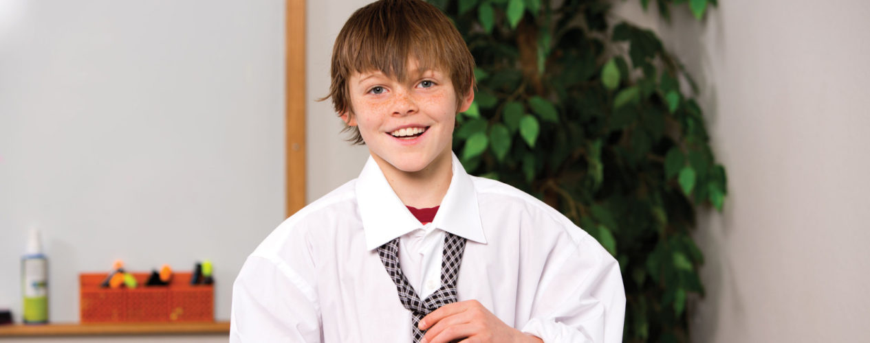 A preteen boy is wearing a large man's dress shirt and is tying a tie around his collar as he celebrates a late spring holiday.