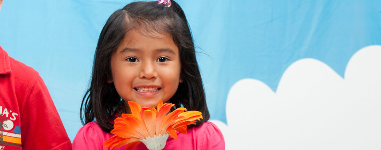 A preschool girl holding an orange flower up to her face. She is standing in front of a blue sky backdrop.