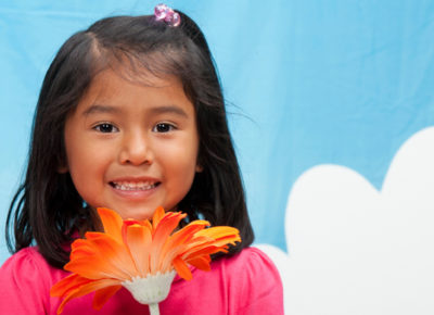 A preschool girl holding an orange flower up to her face. She is standing in front of a blue sky backdrop.