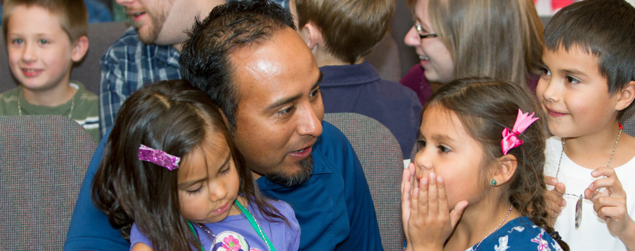 Father sits in a sanctuary with his two daughters and son. One daughter excited has her hands over her mouth.