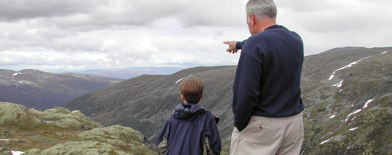 A boy and his grandpa are hiking in a valley. He grandpa is point at something off in the distance.