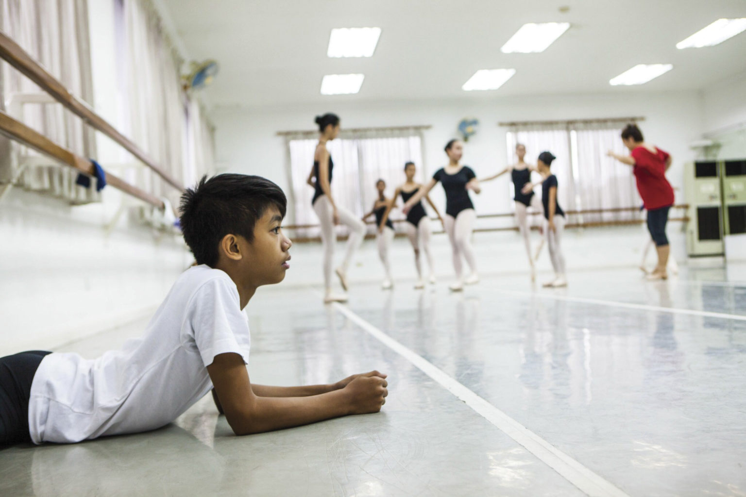A young male dancer lays on his stomach as a ballet class takes place behind him. They are in a studio.