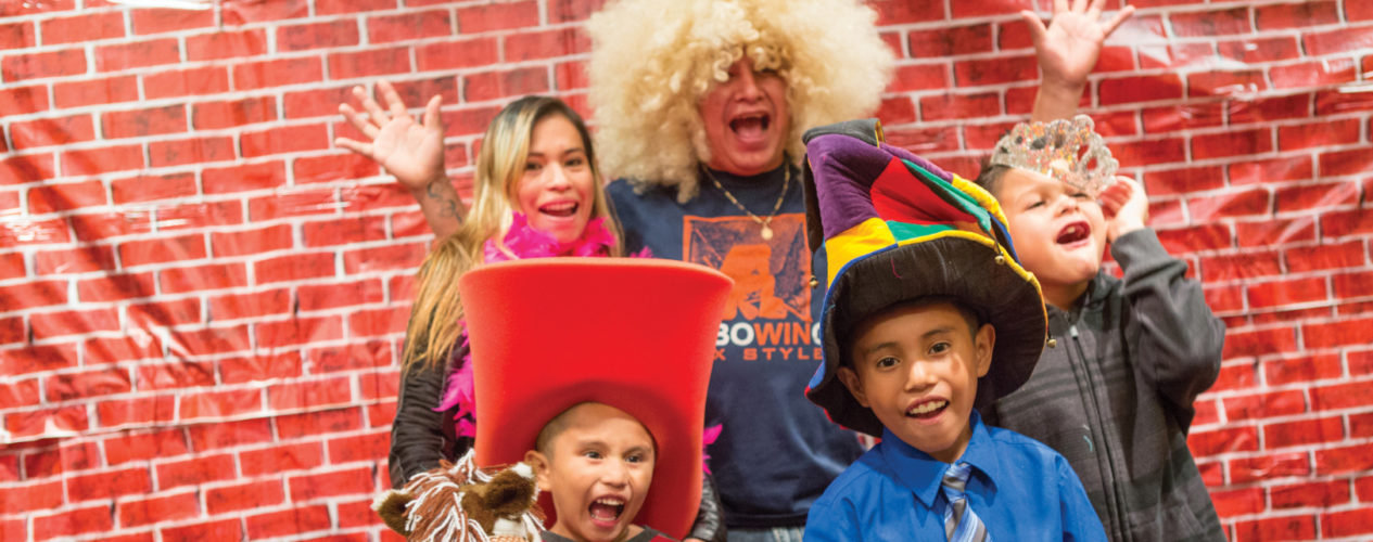 A family wearing goofy props. They are taking a photo in front of a brick wall and making silly faces.