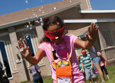 A preteen girl, who is wearing a blindfold, runs through splashing water outside.