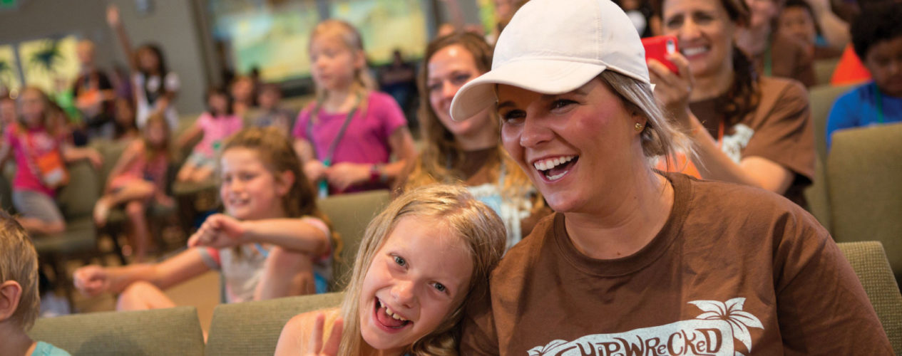 A VBS leader sitting in a sanctuary with an elementary girl sitting next to her. They are both smiling and the girl is making a peace sign with her fingers.