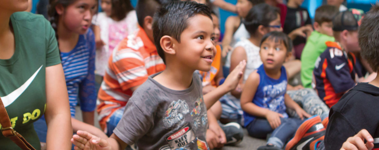 An elementary boy sitting in a crowd of children. He has his arms out at his side participating in some dance moves. He is smiling joyfully at this fall fest, which is used as a VBS follow-up.