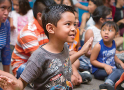 An elementary boy sitting in a crowd of children. He has his arms out at his side participating in some dance moves. He is smiling joyfully at this fall fest, which is used as a VBS follow-up.