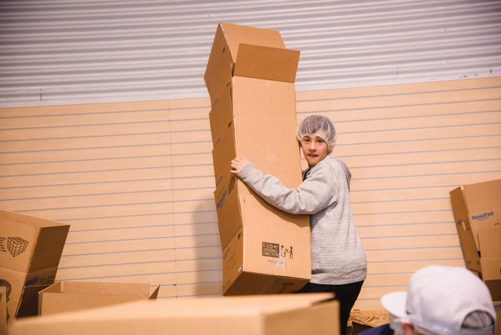 Teenage boy in a hair net and sweatshirt is holding a stack of empty cardboard boxes that are towering over him.