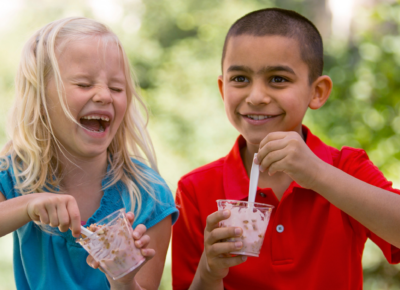 A little girl in a blue shirt is laughing as she eats independence ice cream with a spoon. She is sitting next to a boy in a red shirt eating ice cream as well.