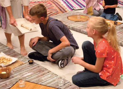 Woman dressed in Biblical clothing bending down to let a boy and girl sitting around a table on the ground smell the food that's on the plate.